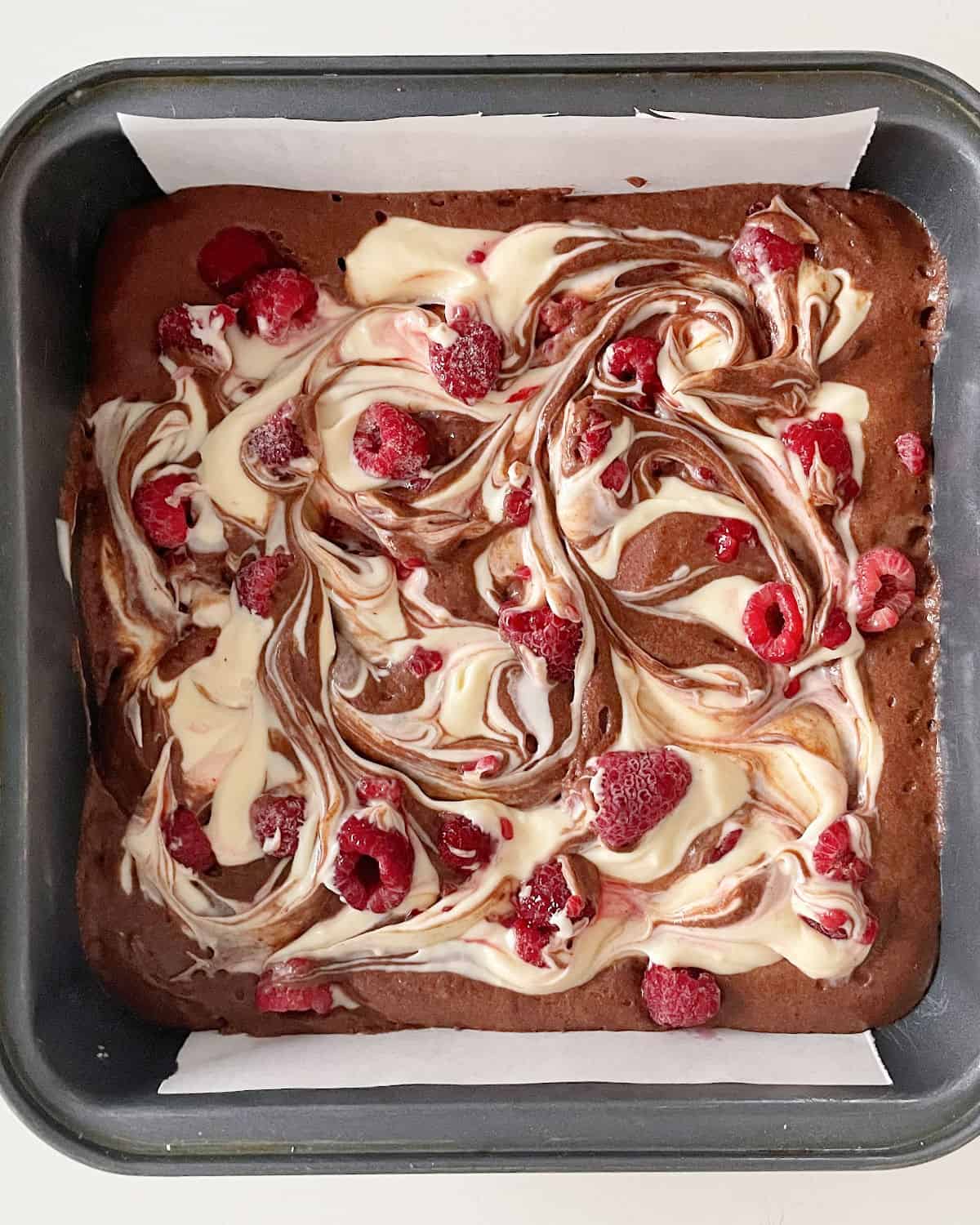 Swirled cheesecake brownie batter with raspberries in a metal square pan. 