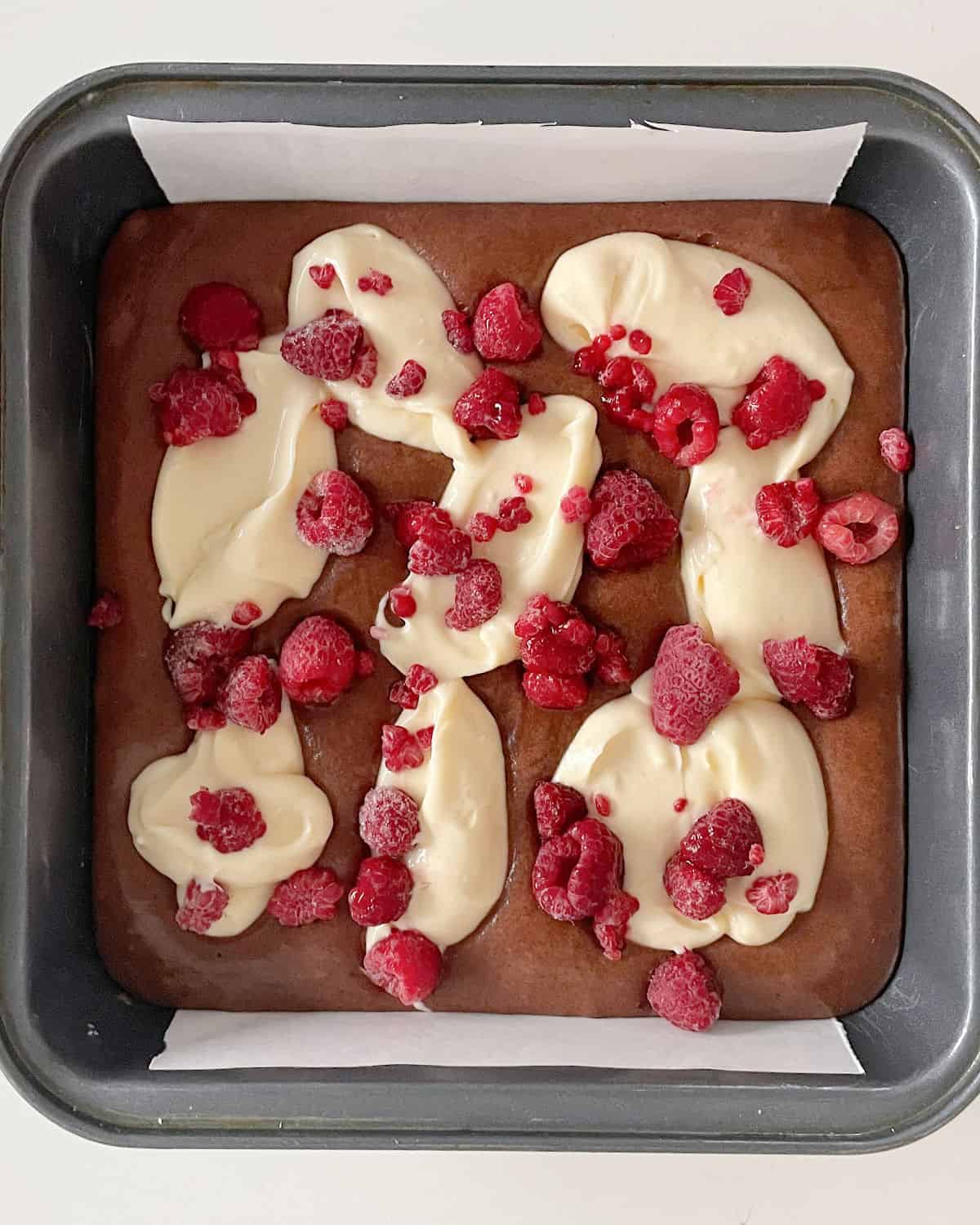 Layered brownie batter, cheesecake mix, and raspberries in a square metal pan on a white surface.