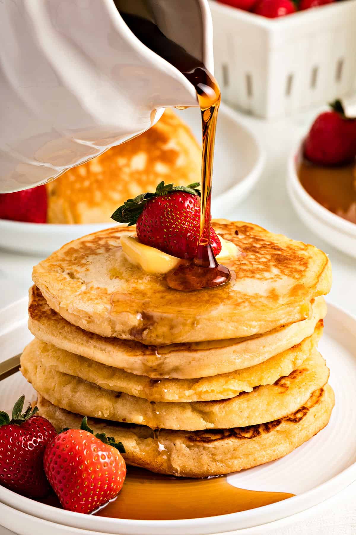 Stack of pancakes with syrup being poured on them. White plate and background.