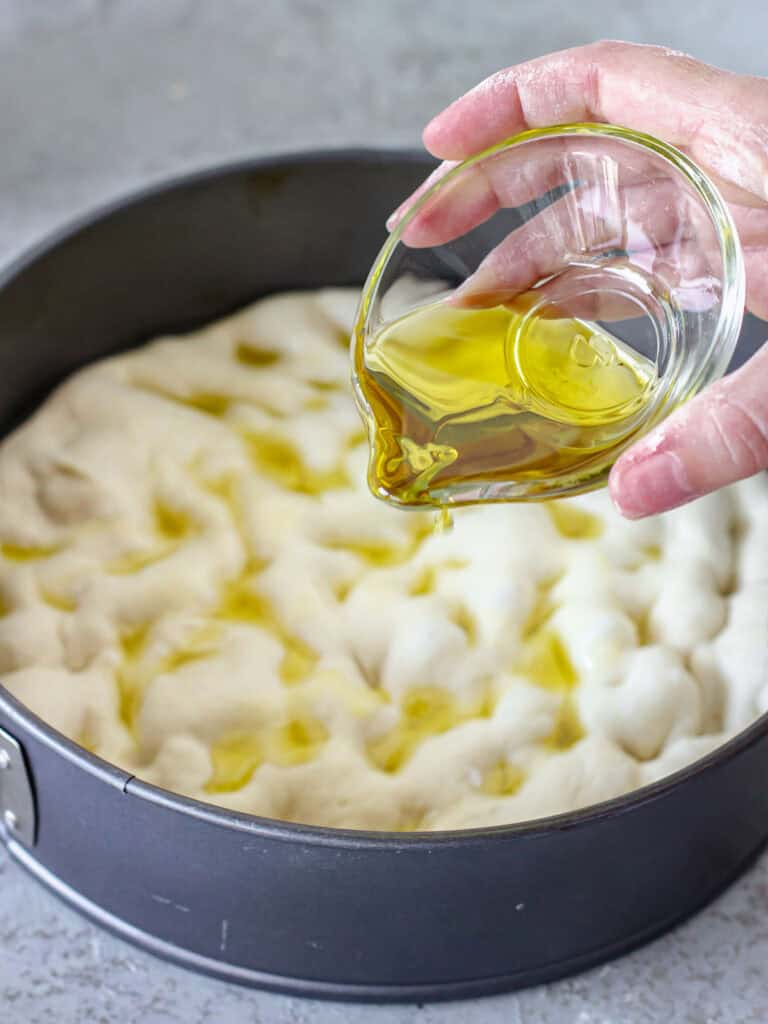 Drizzling oil on dimpled focaccia dough in a round dark metal cake pan.