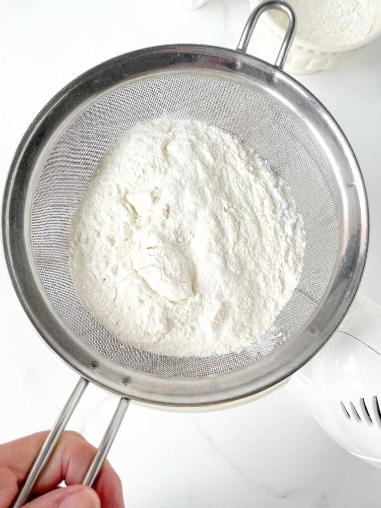 Sifting flour mixture over glass bowl with cake batter. White surface. Top view.