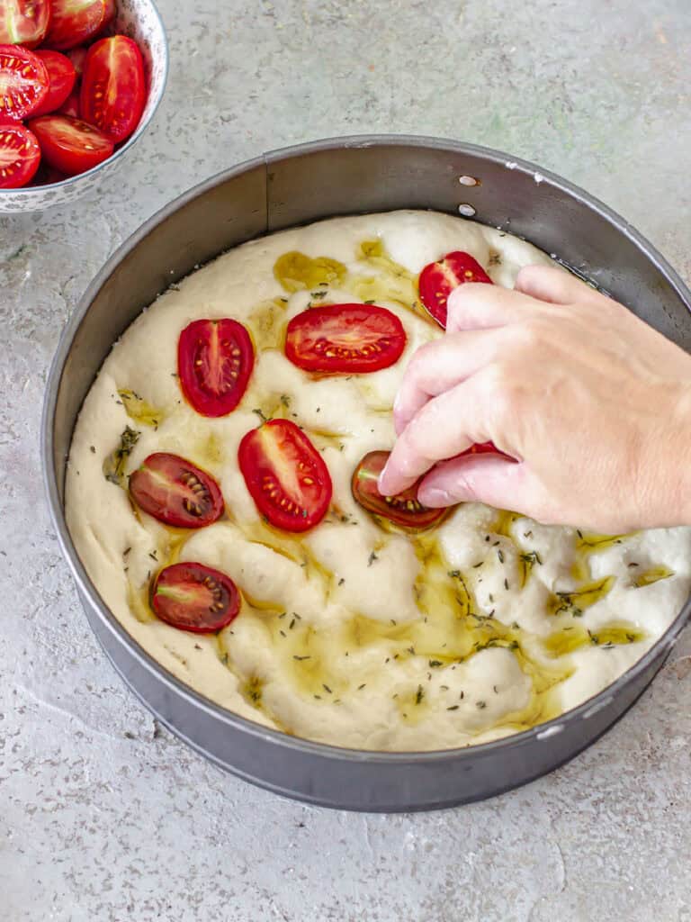 Placing halved cherry tomatoes on focaccia dough in a round pan. Gray background. 