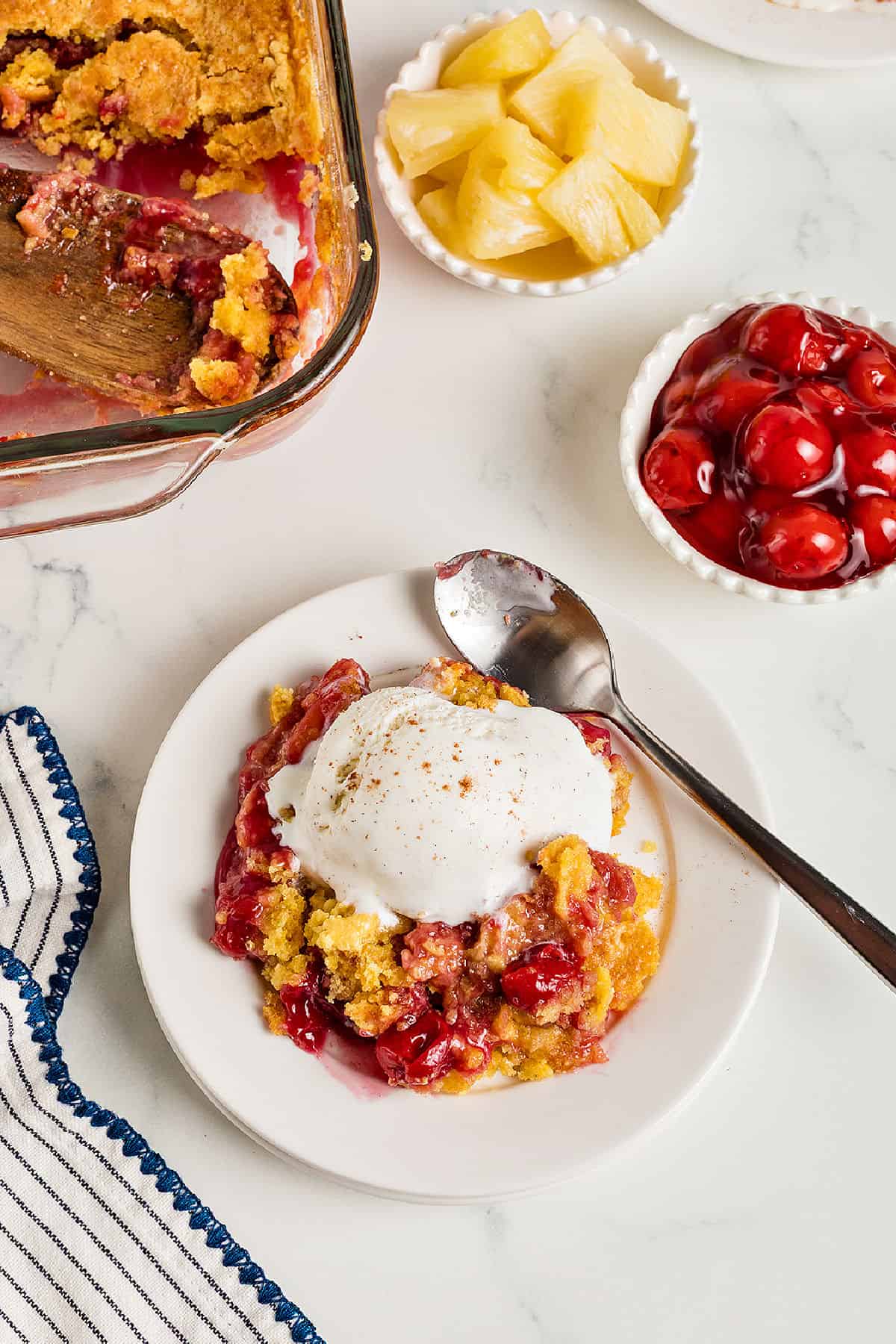 Cherry pineapple cobbler with ice cream and silver spoon. White plate, bowls with fruit, glass dish. White surface.