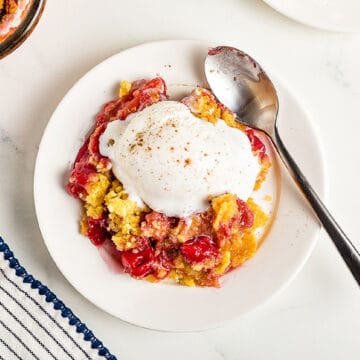 Serving of ice-cream topped cherry pineapple cobbler. Silver spoon, white plate and surface.