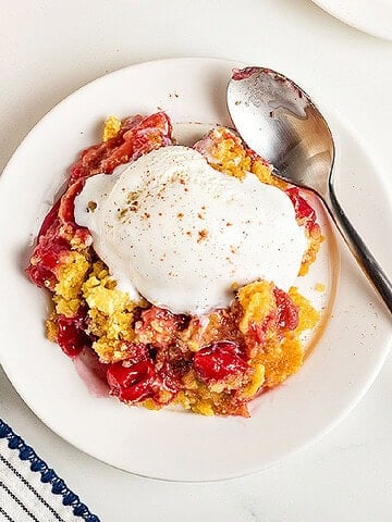 Serving of ice-cream topped cherry pineapple cobbler. Silver spoon, white plate and surface.