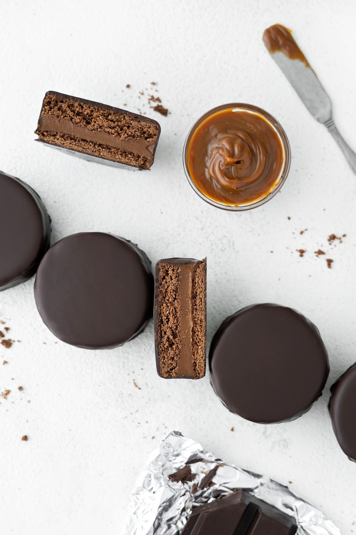 Top view of white surface with halved and whole chocolate alfajores. Bowl of dulce de leche, butter knife.