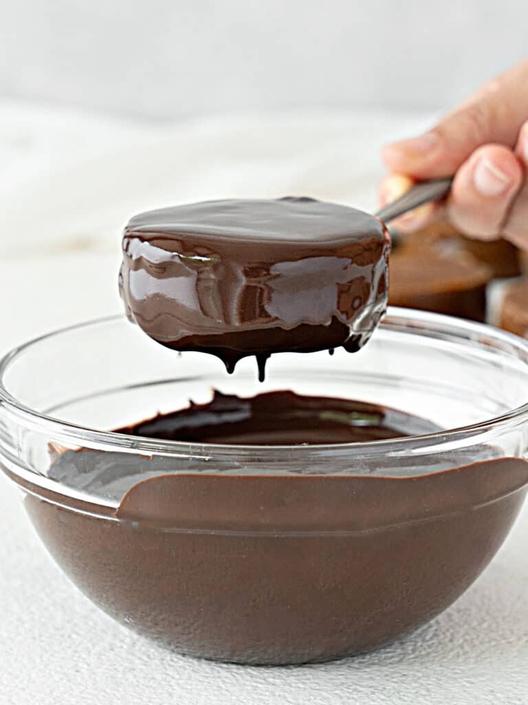 Lifting chocolate covered alfajor from glass bowl of melted chocolate. White background.