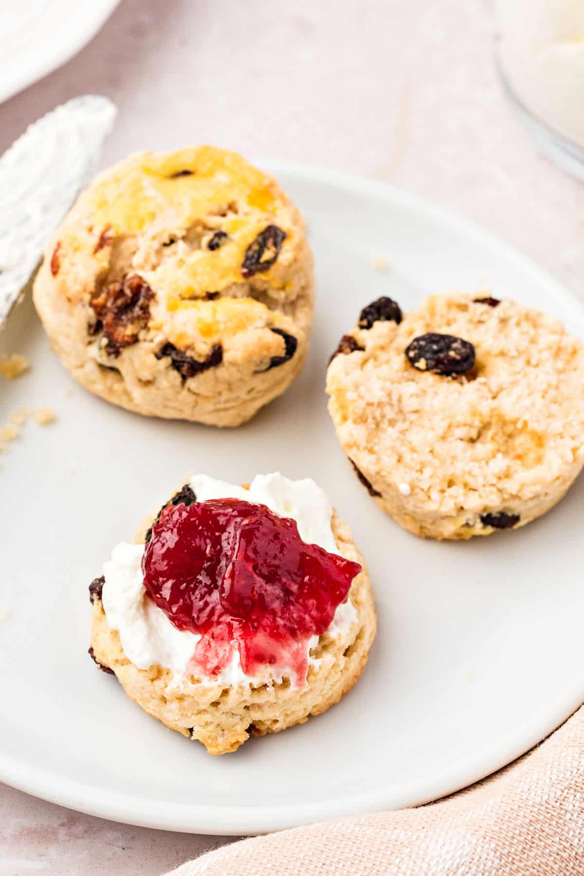 Cream and jam topped scone with whole one on a white plate.