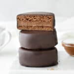 Three stacked chocolate covered alfajores. Top one is halved. White background.