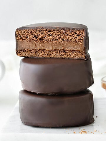 Three stacked chocolate covered alfajores. Top one is halved. White background.