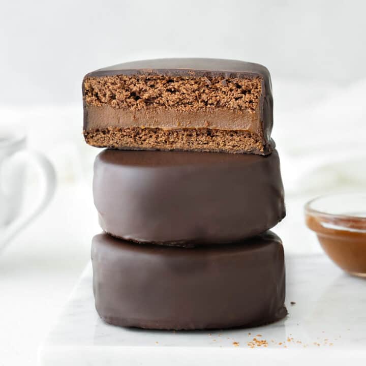 Three stacked chocolate covered alfajores. Top one is halved. White background.