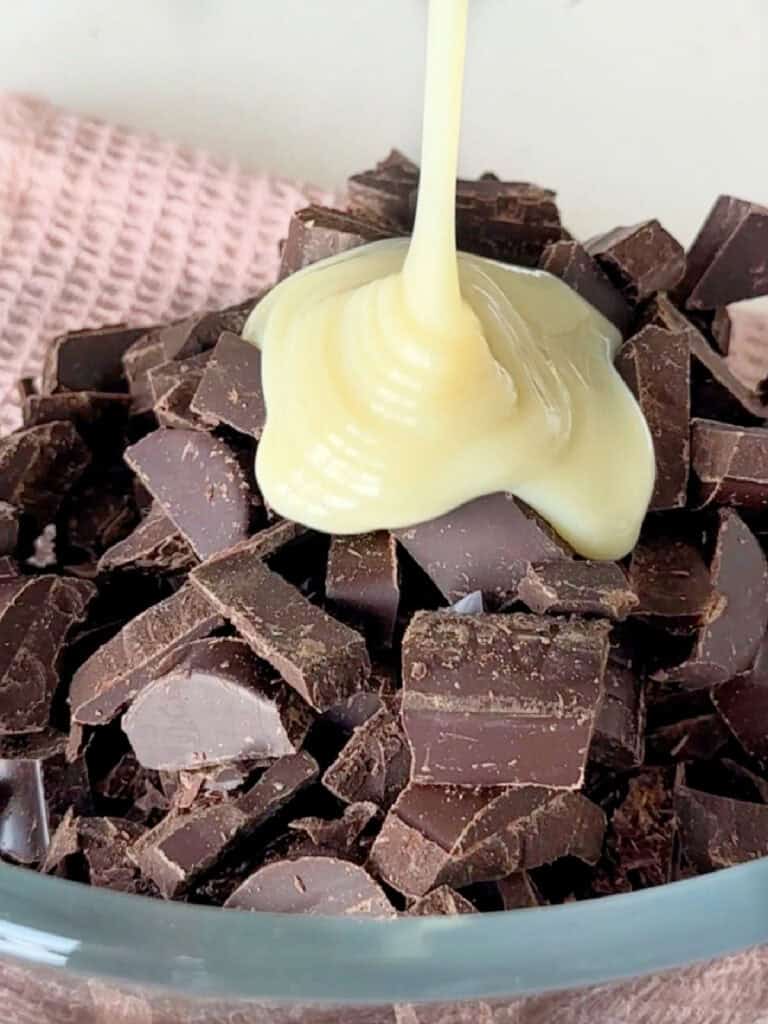 Condensed milk being poured over dark chocolate pieces.