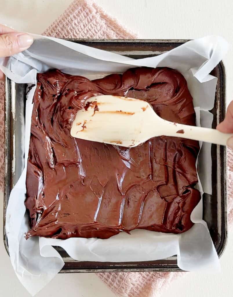 Smoothing top of fudge mixture with a spatula in a paper lined pan.