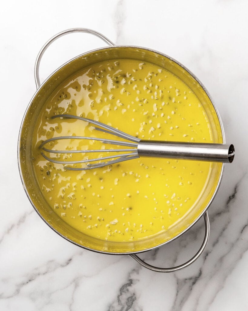 Metal saucepan and whisk with passion fruit curd on white marble.
