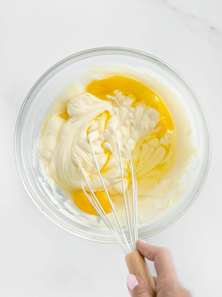 Mixing eggs and cream cheese in a glass bowl with a whisk. White background.