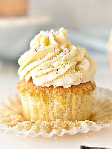 Close up frosted vanilla cupcake with sprinkles. Flat paper liner. White gray background.