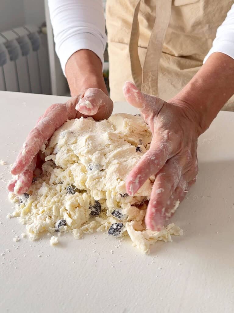 Hands gathering raisin scones dough on a white table.
