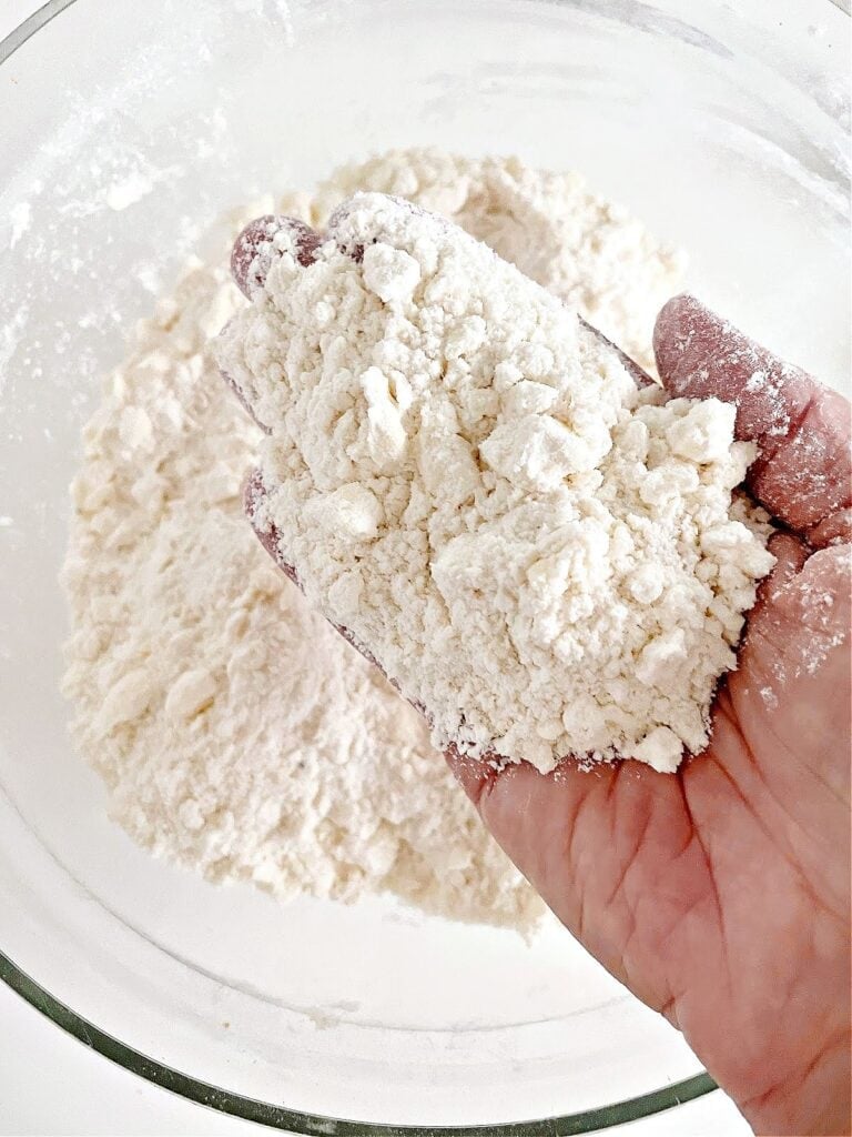 Hand with flour and small butter pieces over a glass bowl.