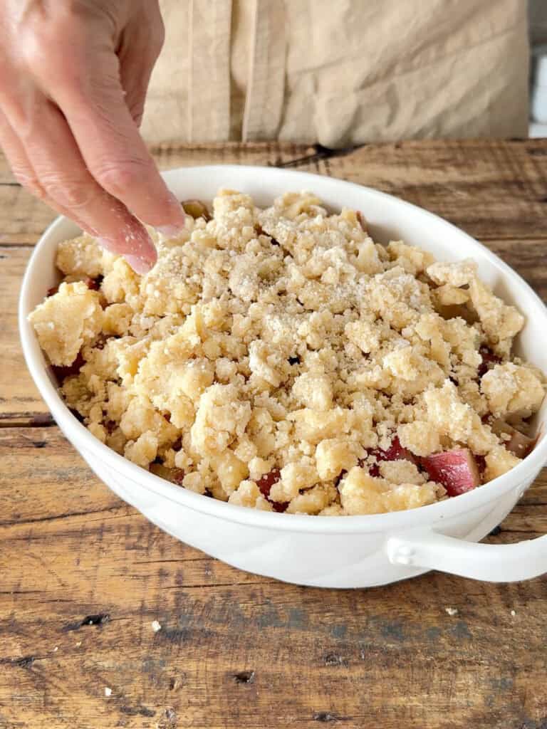 Sprinkling crumble mixture over rhubarb in oval baking dish. Wooden surface.