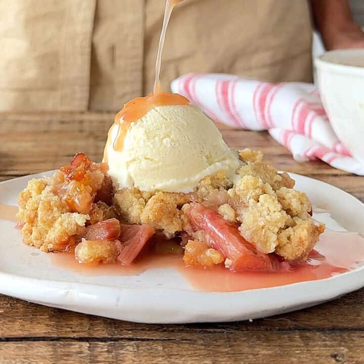 Close up pouring syrup over ice cream and rhubarb crumble on a white plate. Beige background.
