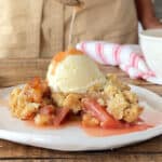 Close up pouring syrup over ice cream and rhubarb crumble on a white plate. Beige background.
