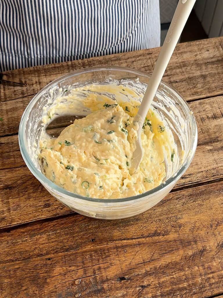 Glass bowl with cheese onion muffin batter and spatula. Wooden table.