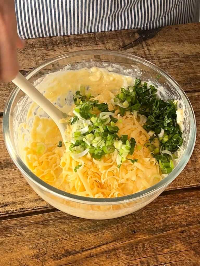 Grated cheese and chopped green onions added to muffin batter in a glass bowl. Spatula. Wooden surface.
