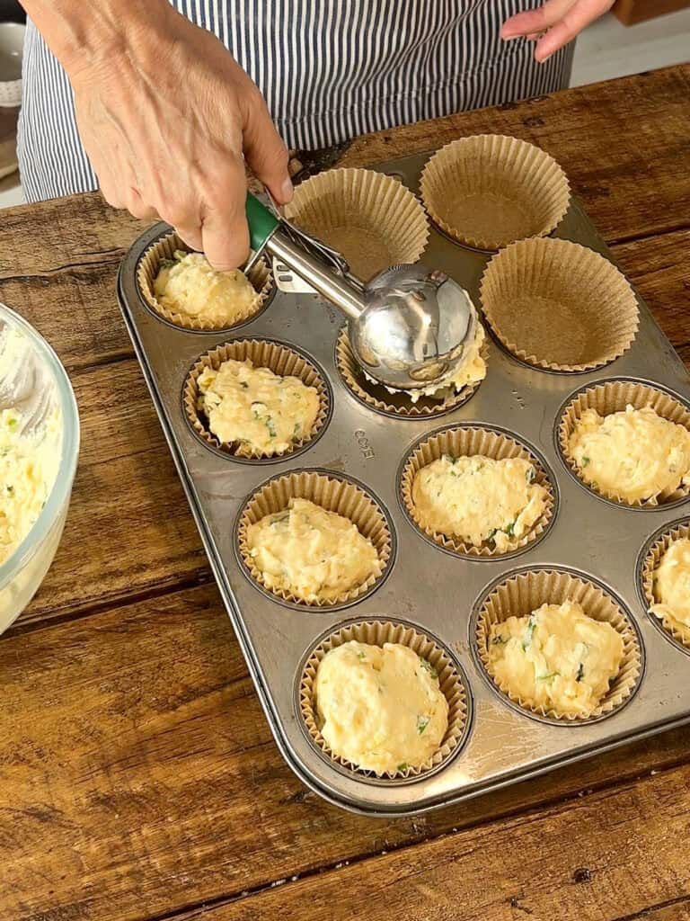 Scooping cheese muffin batter onto beige paper cups in a muffin tin. Wooden table.