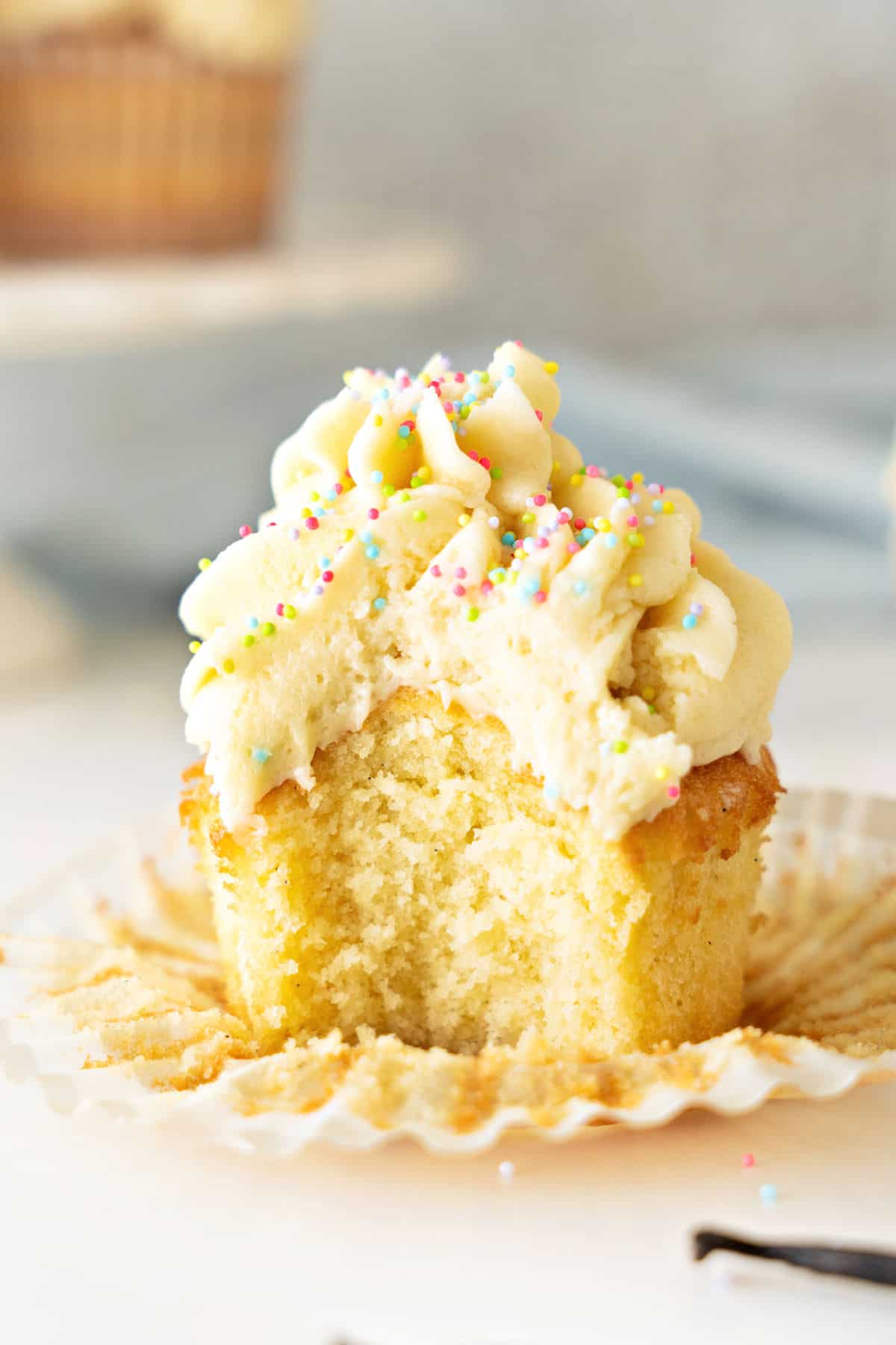 Bitten frosted vanilla cupcake on a flattened paper liner. Gray white background.