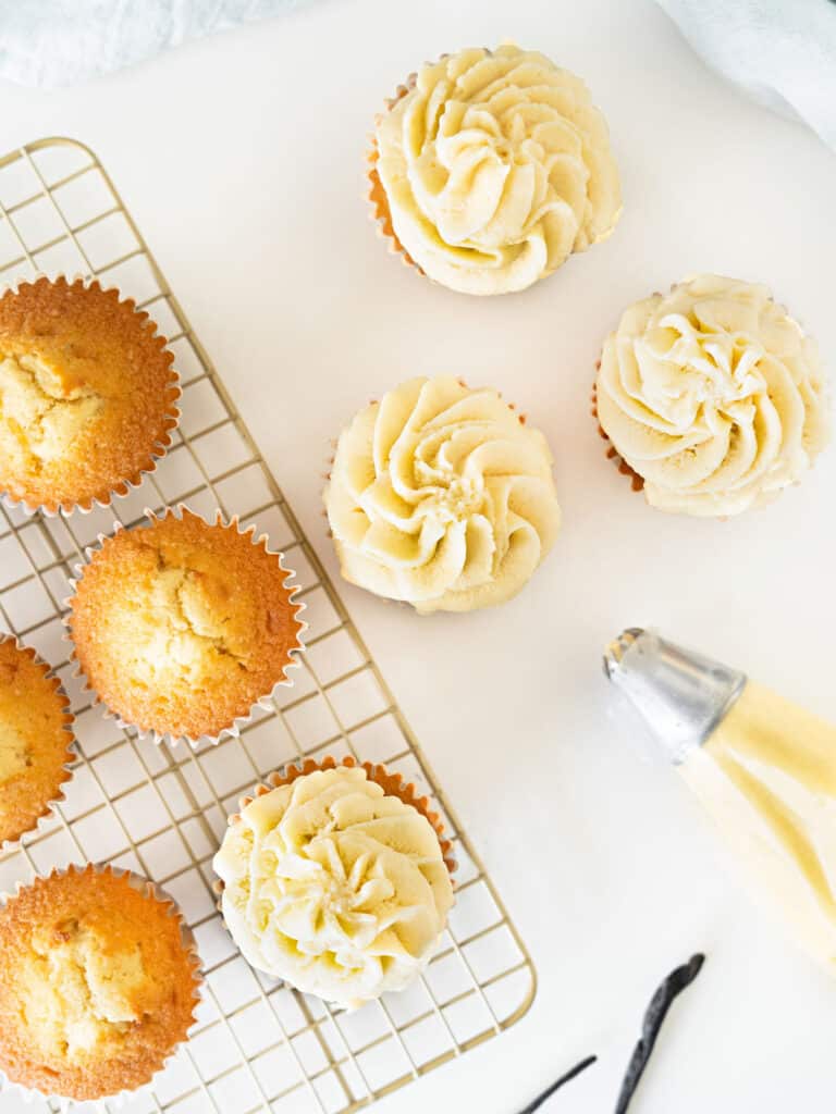 Wire rack with frosted and plain vanilla cupcakes. Piping bag. White surface.