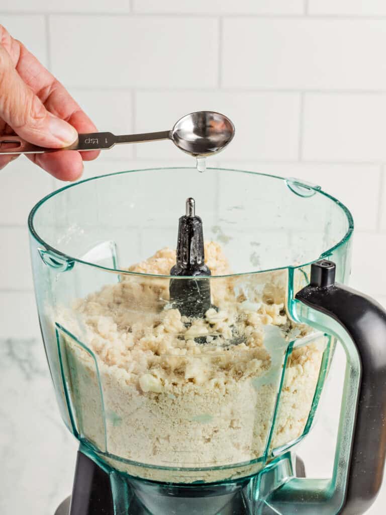Adding spoon of water to pie crust in the food processor bowl.