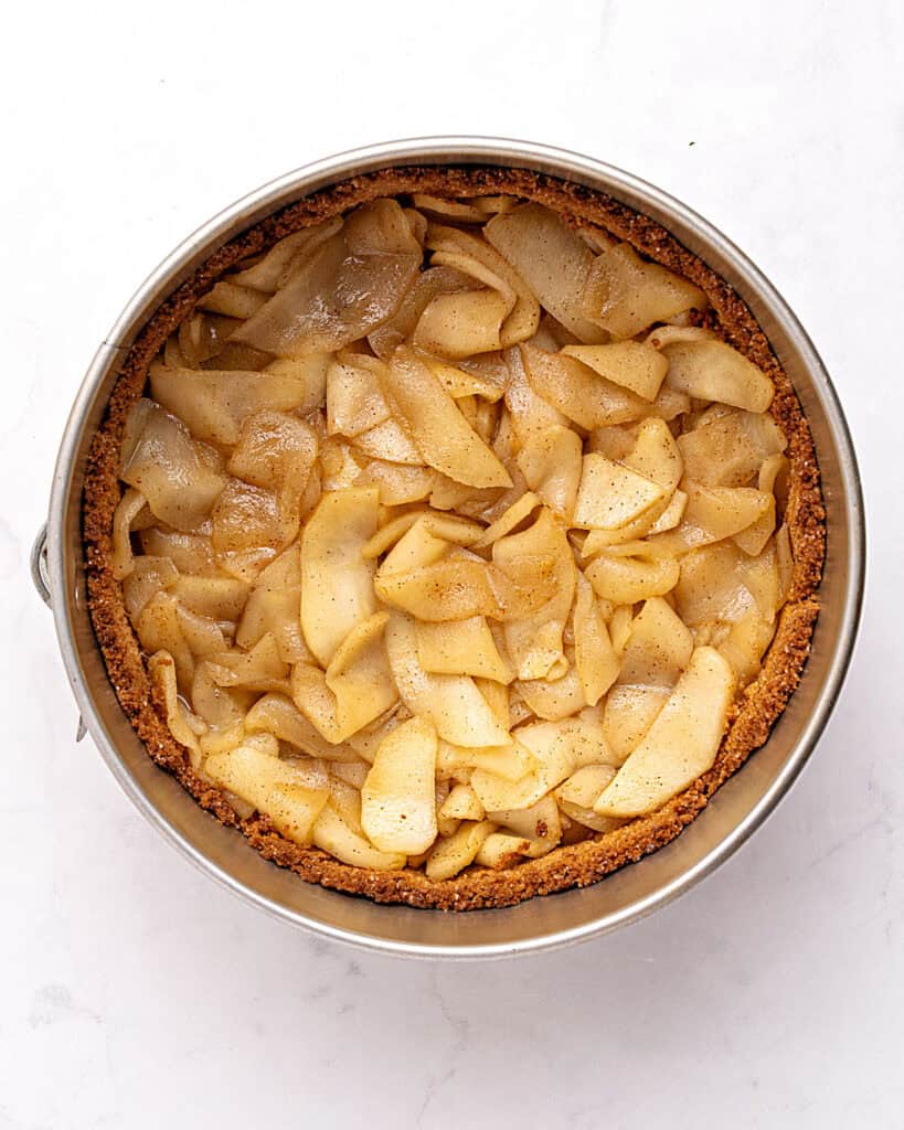Cooked apple layer over a cheesecake crust in a metal cake pan. White surface.