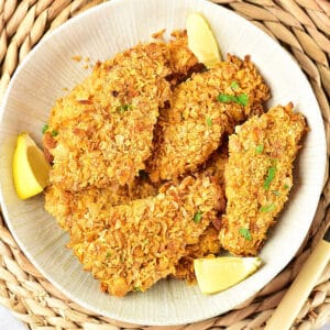 Close up white shallow plate of cornflake chicken breasts and lemon wedges. Raffia underplate.