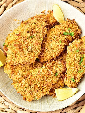 Close up white shallow plate of cornflake chicken breasts and lemon wedges. Raffia underplate.
