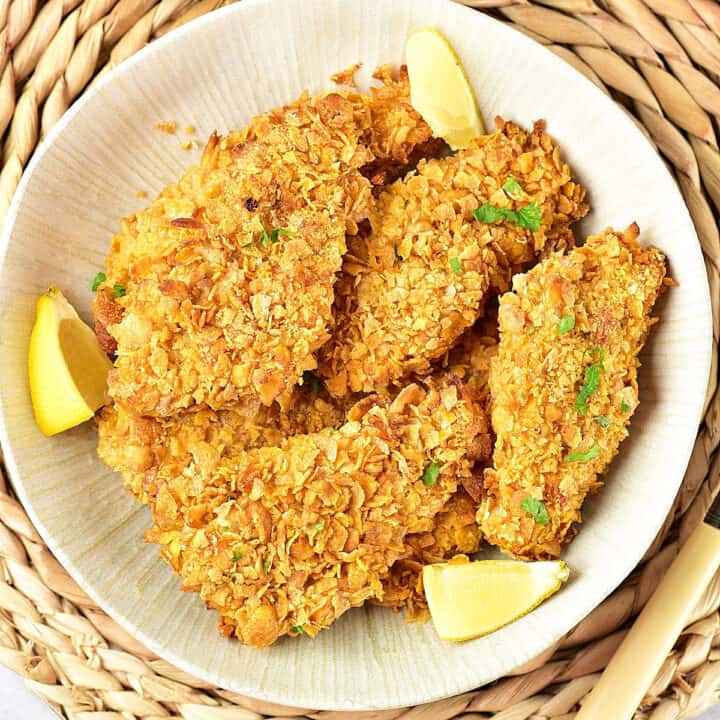 Close up white shallow plate of cornflake chicken breasts and lemon wedges. Raffia underplate.