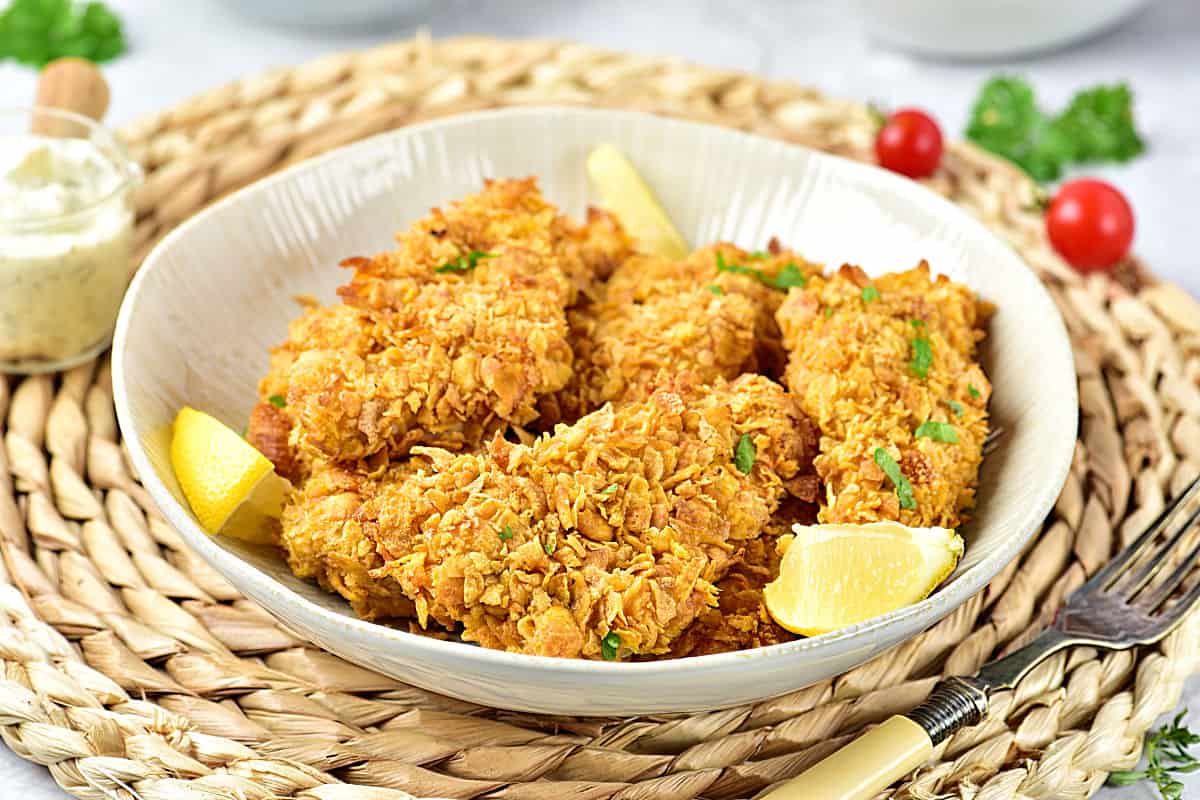 White shallow bowl plate with cornflake chicken tenders. Raffia underplate, lemon wedges.
