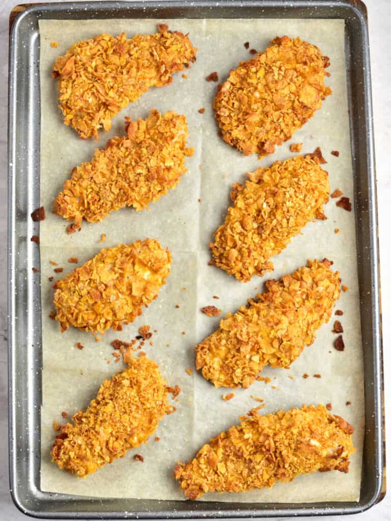 Metal baking sheet with parchment paper and baked cornflake crusted chicken tenders.