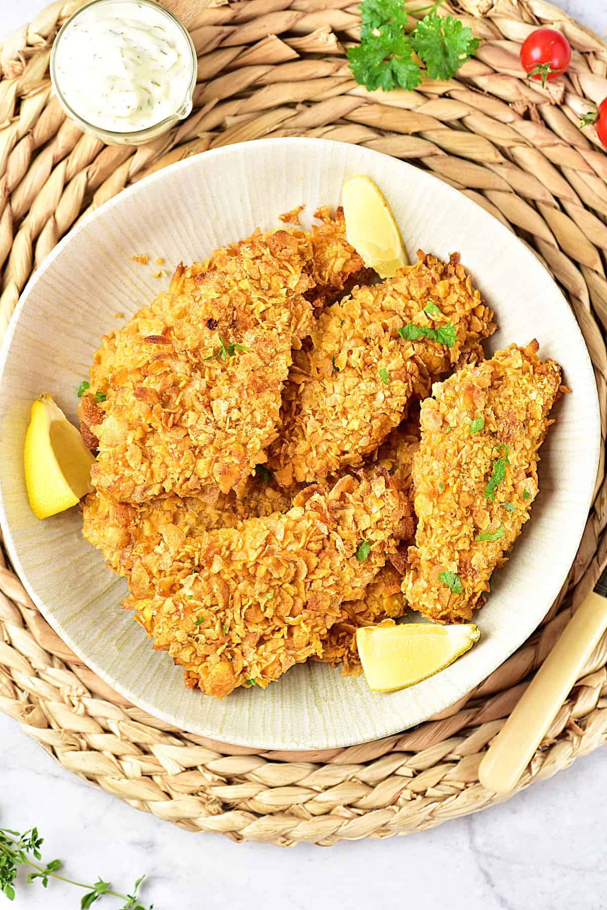 Raffia underplate with white shallow plate of cornflake chicken breasts and lemon wedges.