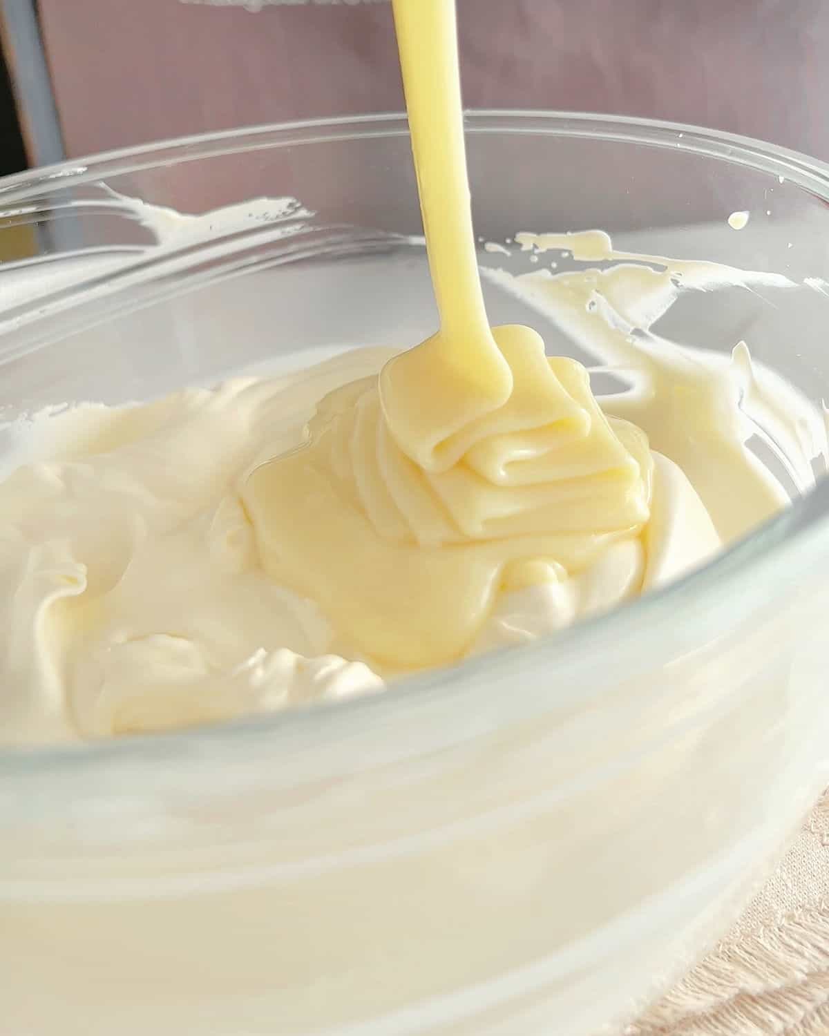 Pouring condensed milk on whipped cream in a glass bowl.