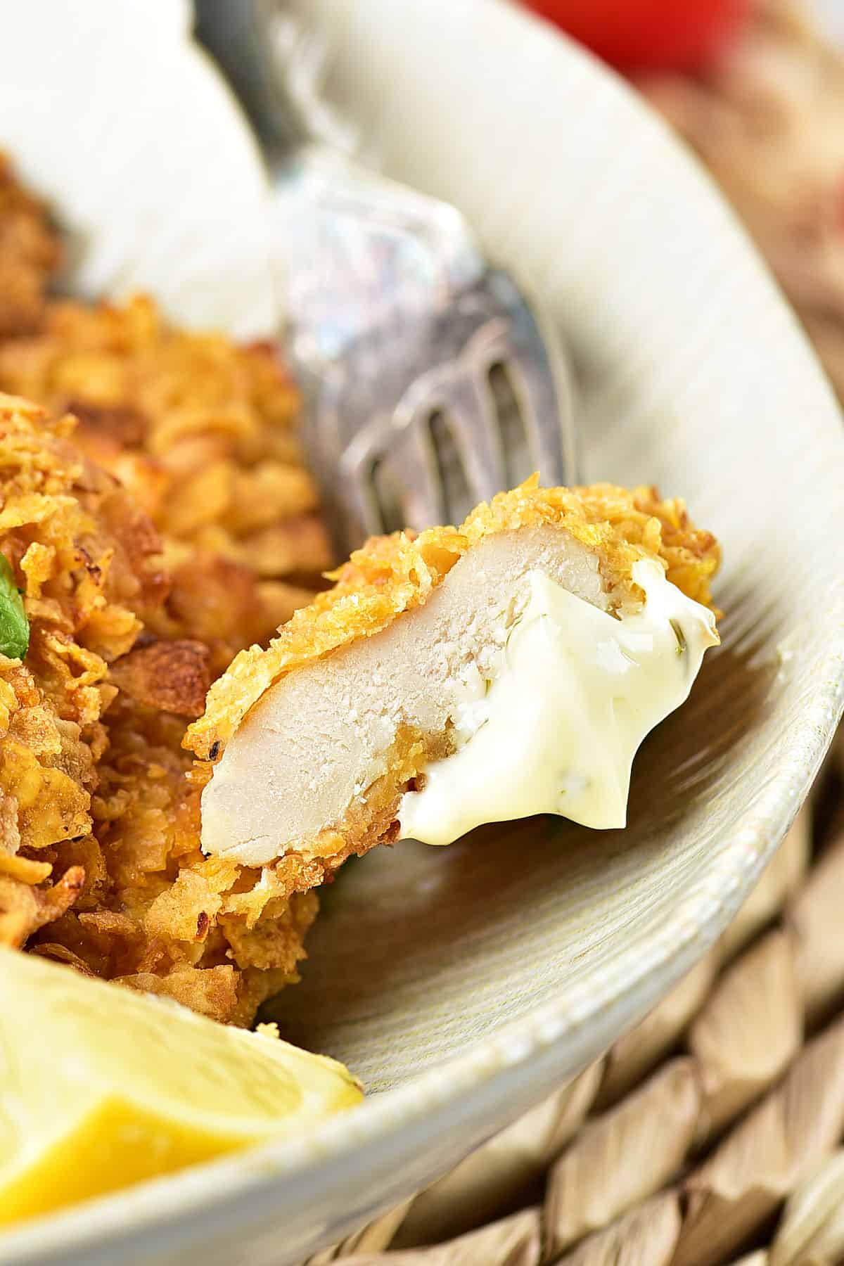 Forked bite of baked cornflake chicken. White plate, lemon wedge.