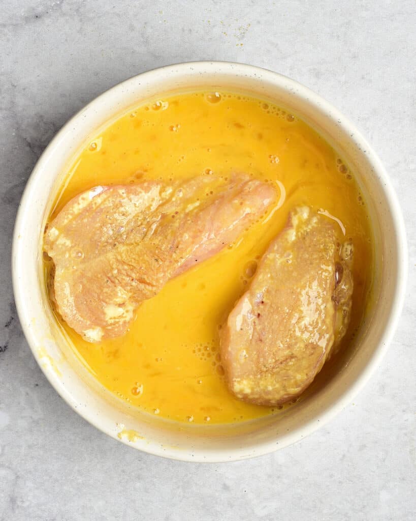 White bowl with egg dipping mixture and chicken breast pieces. Light gray background.
