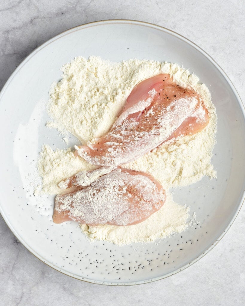 Flour in a shallow plate with chicken breast pieces. Light gray background.