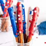 Plastic cup with red, white, blue chocolate-covered pretzel rods with sprinkles. White background.