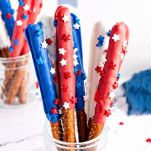 Plastic cup with red, white, blue chocolate-covered pretzel rods with sprinkles. White background.