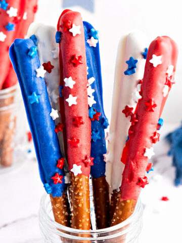 Plastic cup with red, white, blue chocolate-covered pretzel rods with sprinkles. White background.
