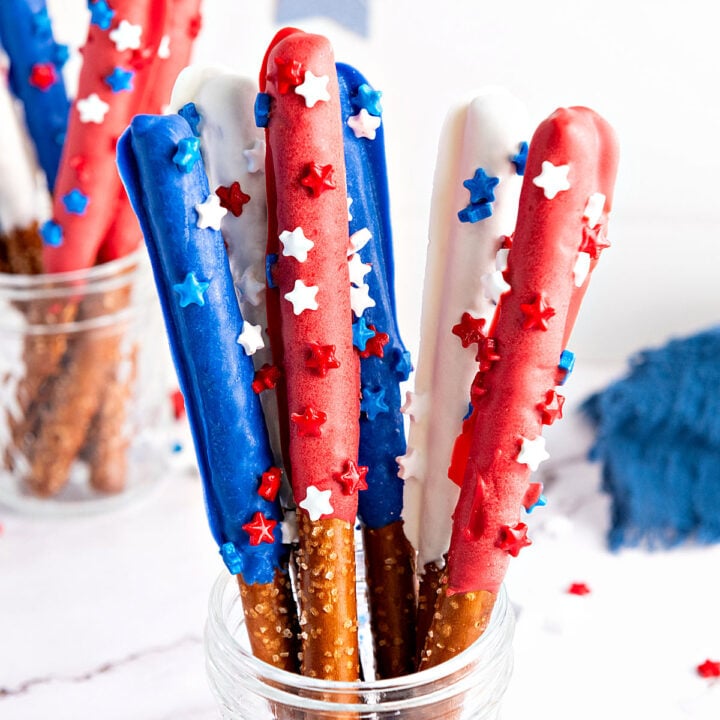 Plastic cup with red, white, blue chocolate-covered pretzel rods with sprinkles. White background.