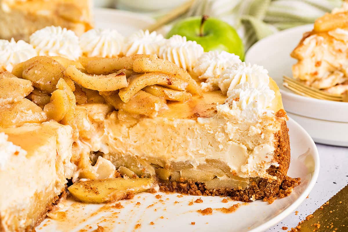 Cut whole apple cheesecake on a white plate with cream and apple topping.