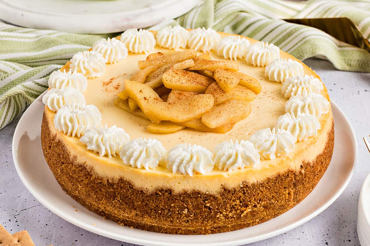 Whole apple cheesecake on a white plate. Green towel in the background.