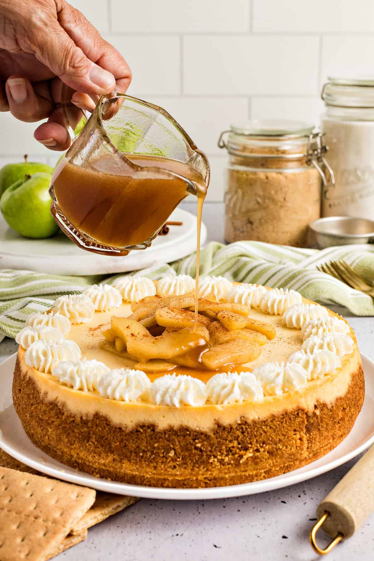 Cream and apple topped cheesecake having syrup poured on. Apples, green cloth, brown sugar jar, tile background.