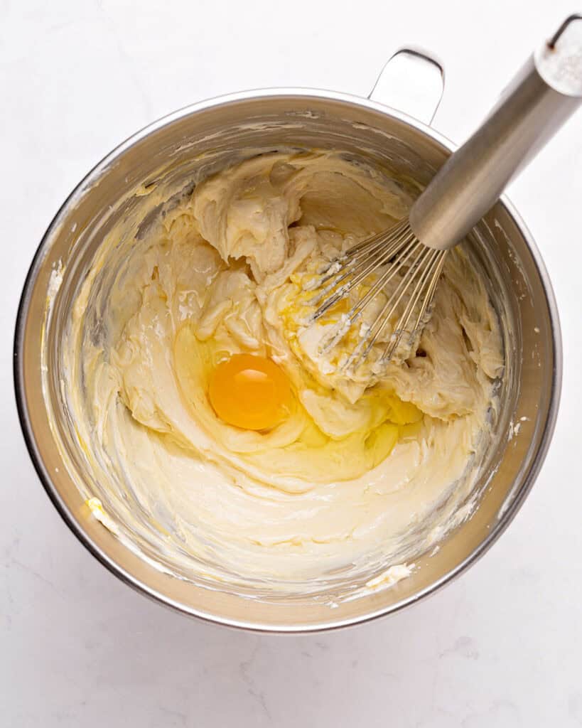 Egg and cheesecake filling in a metal bowl with a whisk. Light gray background.